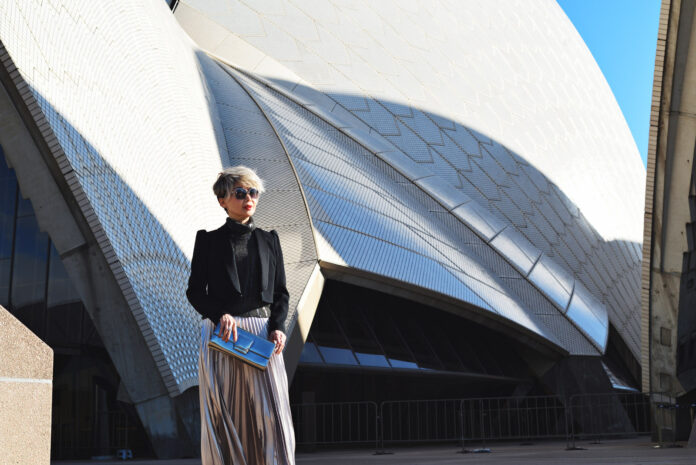 A portrait of writer and blogger Vivienne She at the Sydney Opera House. Vivienne wears a charcoal knitted top with a satin lapel black blazer and a silver pleated midi skirt. Fashion portrait by Sydney photographer Kent Johnson for White Caviar Life.