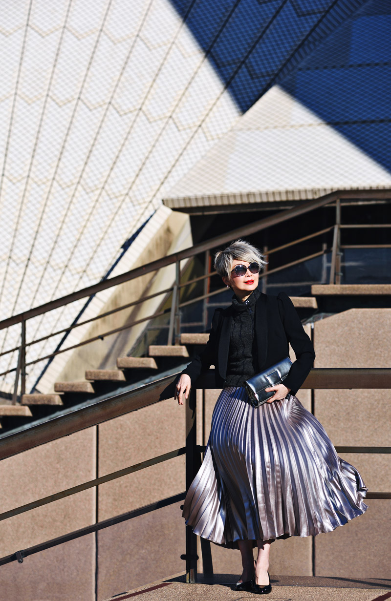 A portrait of writer and blogger Vivienne She at the Sydney Opera House. Vivienne wears a charcoal knitted top with a satin lapel black blazer and a silver pleated midi skirt. Fashion portrait by Sydney photographer Kent Johnson for White Caviar Life.